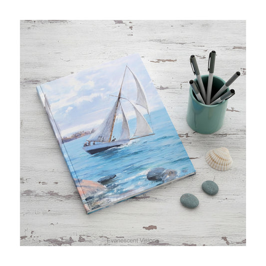 A hardcover notebook depicting a white sailboat sailing on blue water under a cloudy sky, from painting 'Sailing' by Danielson-Gambogi, shown on a distressed white desk with shells and pebbles.