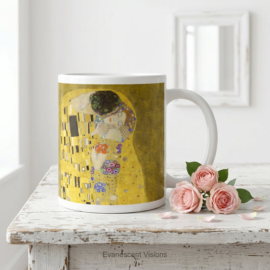 A white ceramic mug featuring a print of Gustav Klimt's 'The Kiss' painting shown on a white wood table with small pink roses.