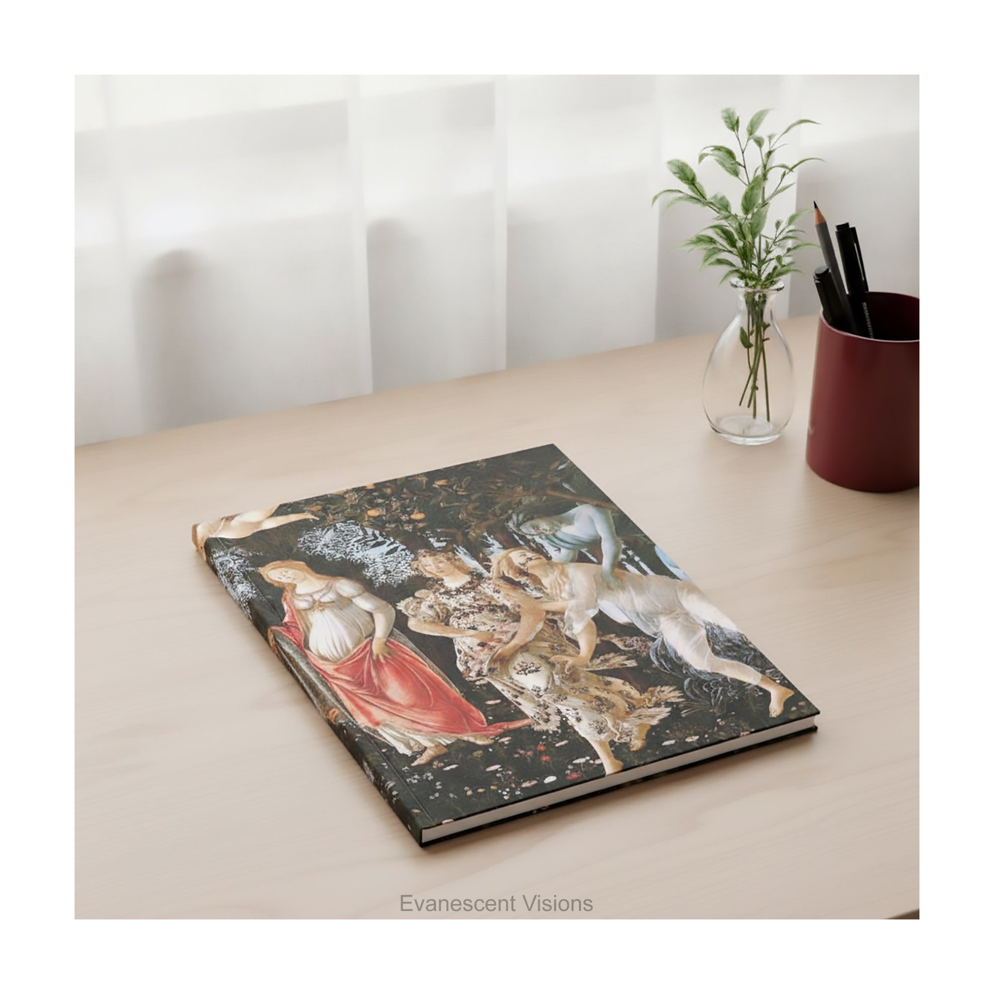 A hardcover notebook featuring the classical painting titled 'Primavera' by Sandro Boticelli, shown lying on a wood desk with pen pot and a small vase. 