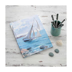 A hardcover notebook  depicting a white sailboat sailing on blue water under a cloudy sky, from painting 'Sailing' by Danielson-Gambogi, shown on a distressed white desk with shells and pebbles. 