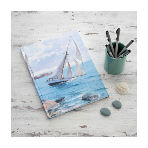 A hardcover notebook  depicting a white sailboat sailing on blue water under a cloudy sky, from painting 'Sailing' by Danielson-Gambogi, shown on a distressed white desk with shells and pebbles. 