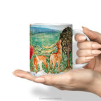 Hands holding a ceramic mug with Pre-Raphaelite painting 'Deer by a Fountain'.