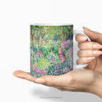 Hands holding a ceramic mug with a print of artwork The Artist's Garden at Giverney' by Claude Monet