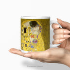 A hand holding a white ceramic mug featuring a print of  Gustav Klimt's 'The Kiss' painting shown on a white background.