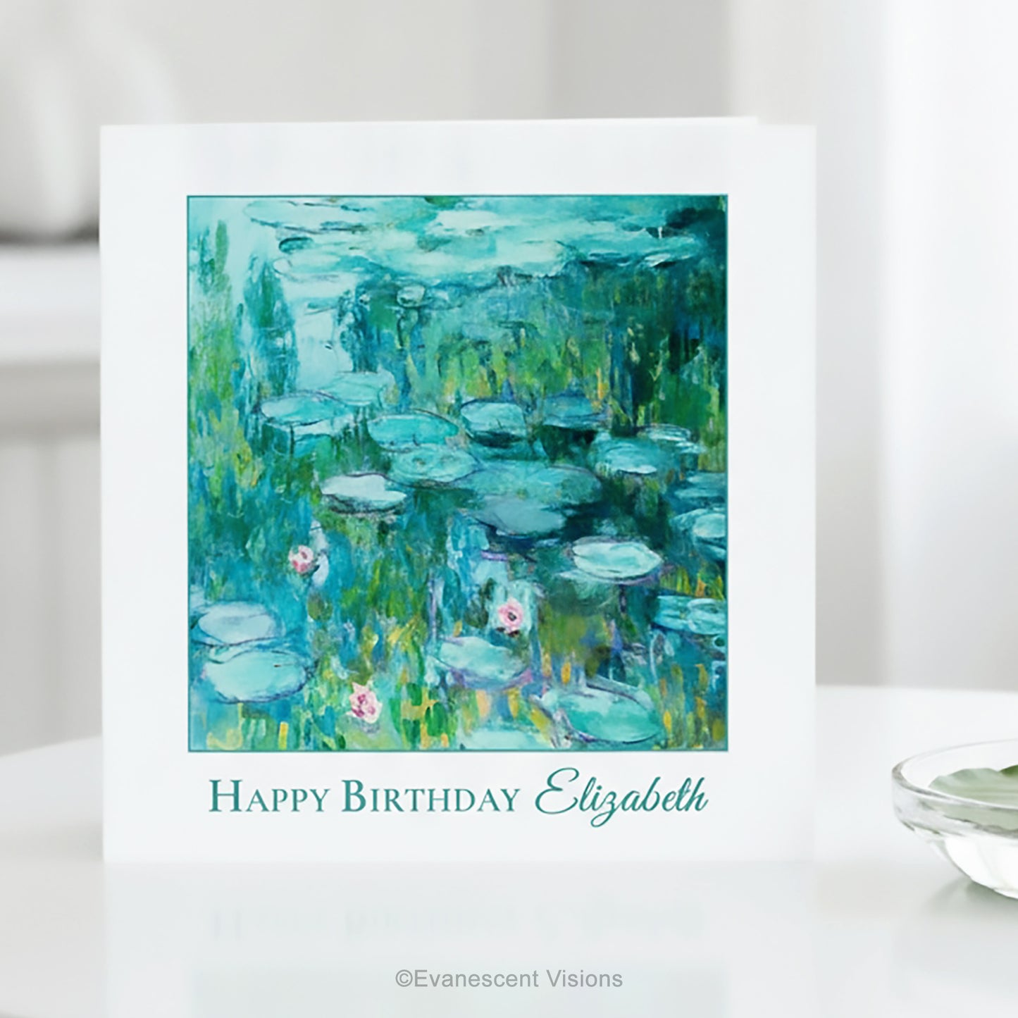 A white greeting card standing on a white surface, featuring Claude Monet's impressionist Water Lilies Painting in blue, green, and teal tones with the text 'Happy Birthday Elizabeth' below the artwork. 