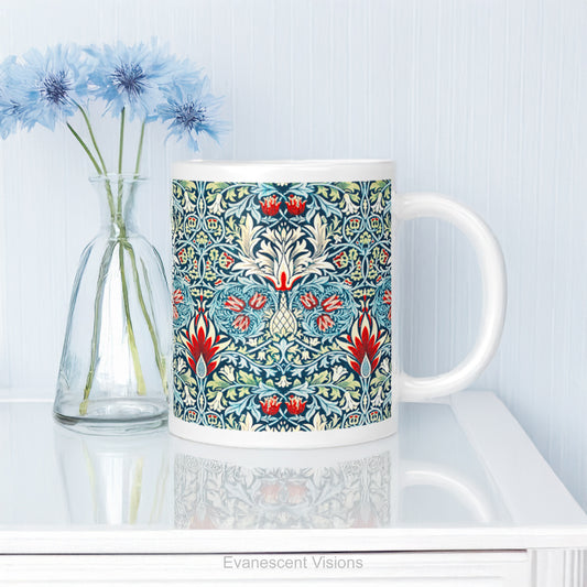 A white ceramic mug featuring a detailed, symmetrical floral pattern in navy blue, sage green, and red called Sankeshead by William Morris,   shown on a glass table top. 