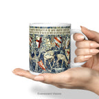 A hand holding a white Ceramic mug with a print of tapestry design 'Verdure with Shields and Deer', shown on a white background.