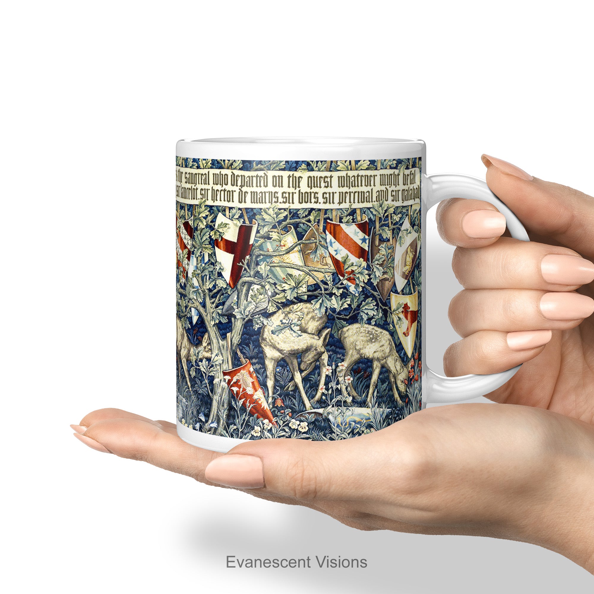 A hand holding a white Ceramic mug with a print of tapestry design 'Verdure with Shields and Deer', shown on a white background.